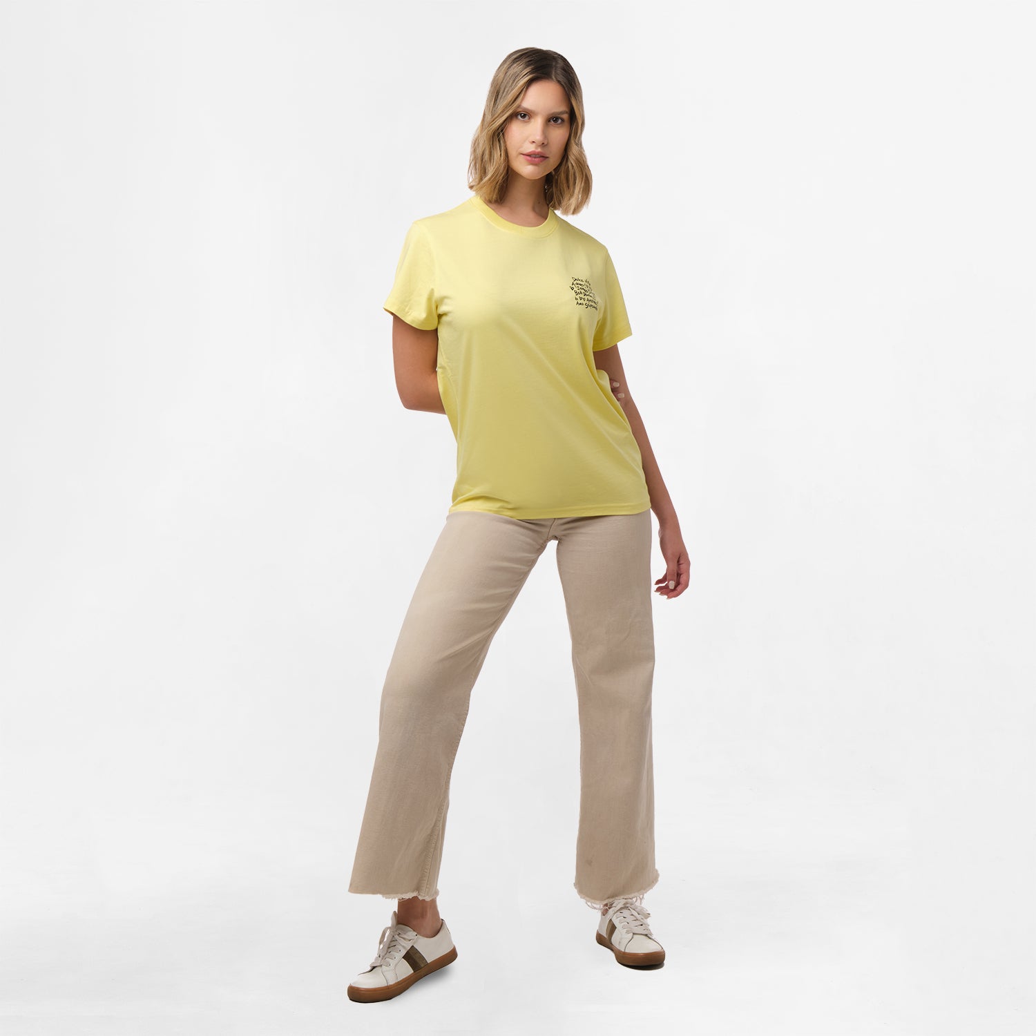 A full-body view of a woman wearing the yellow T-shirt with the black text design on the chest, paired with beige pants, standing with one hand on her hip.