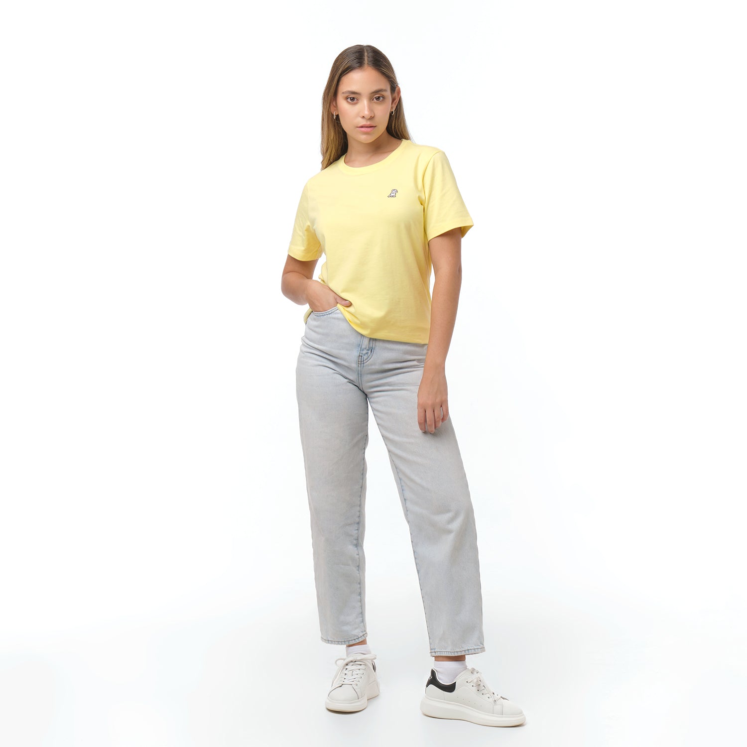 Woman standing full length, posing with hand in pocket, wearing a pale yellow t-shirt with white logo, light wash jeans, and white sneakers