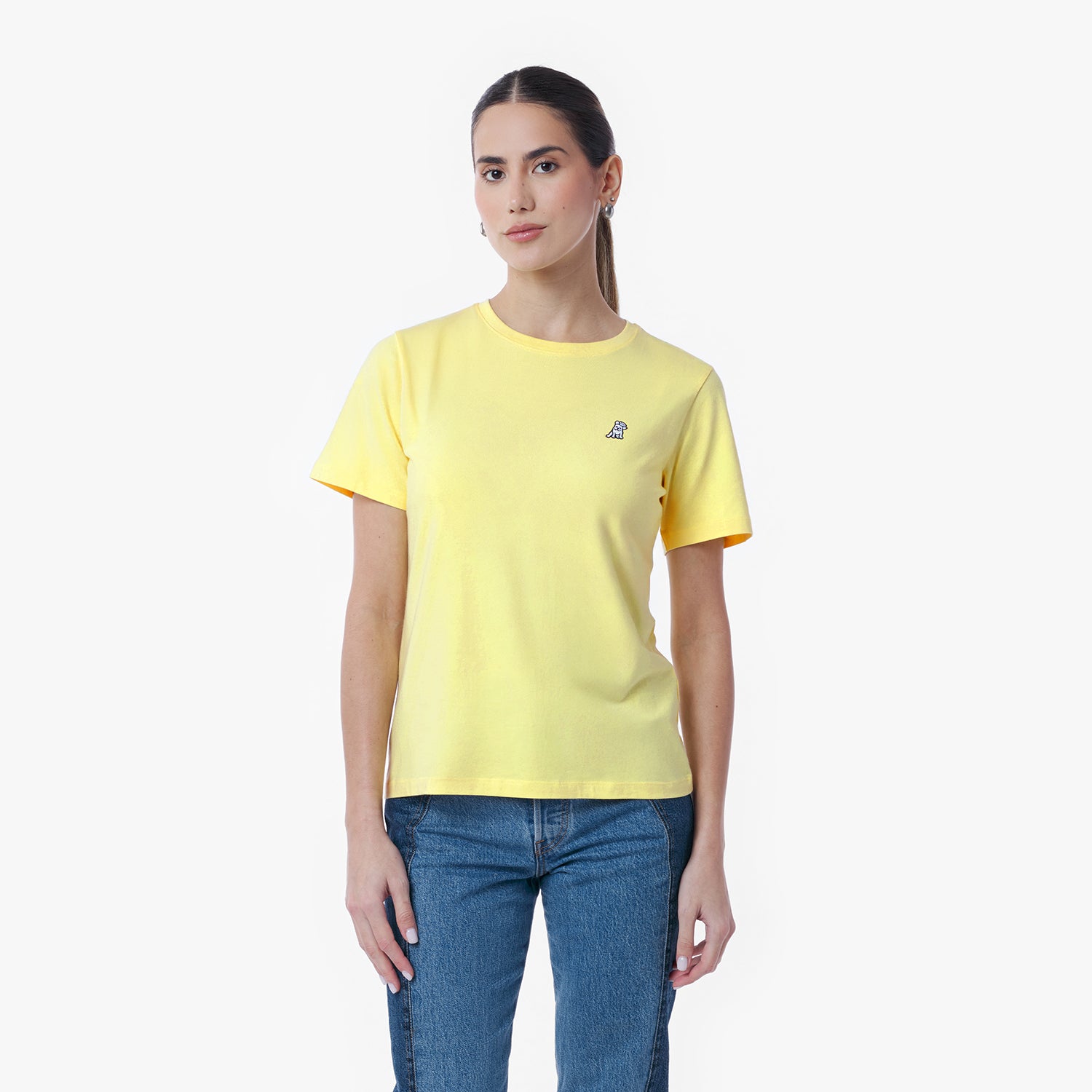 Front view of a woman wearing a yellow crew neck t-shirt with a small logo, blue jeans, and white sneakers, standing against a white background.