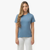 Woman facing forward wearing a dusty blue t-shirt with small white embroidered dog logo on left chest and white wide-leg pants