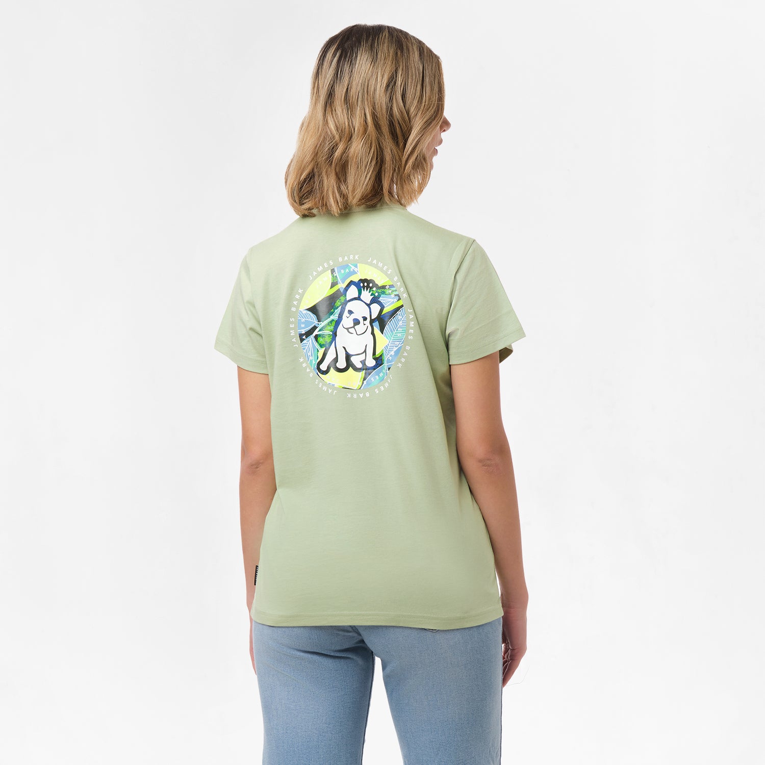 A woman is shown from behind, wearing a light green T-shirt with a large circular graphic on the back. The design features a dog sitting in front of a tropical background with the text "James Bark Privé Society" around the circle. She pairs the shirt with light blue jeans.