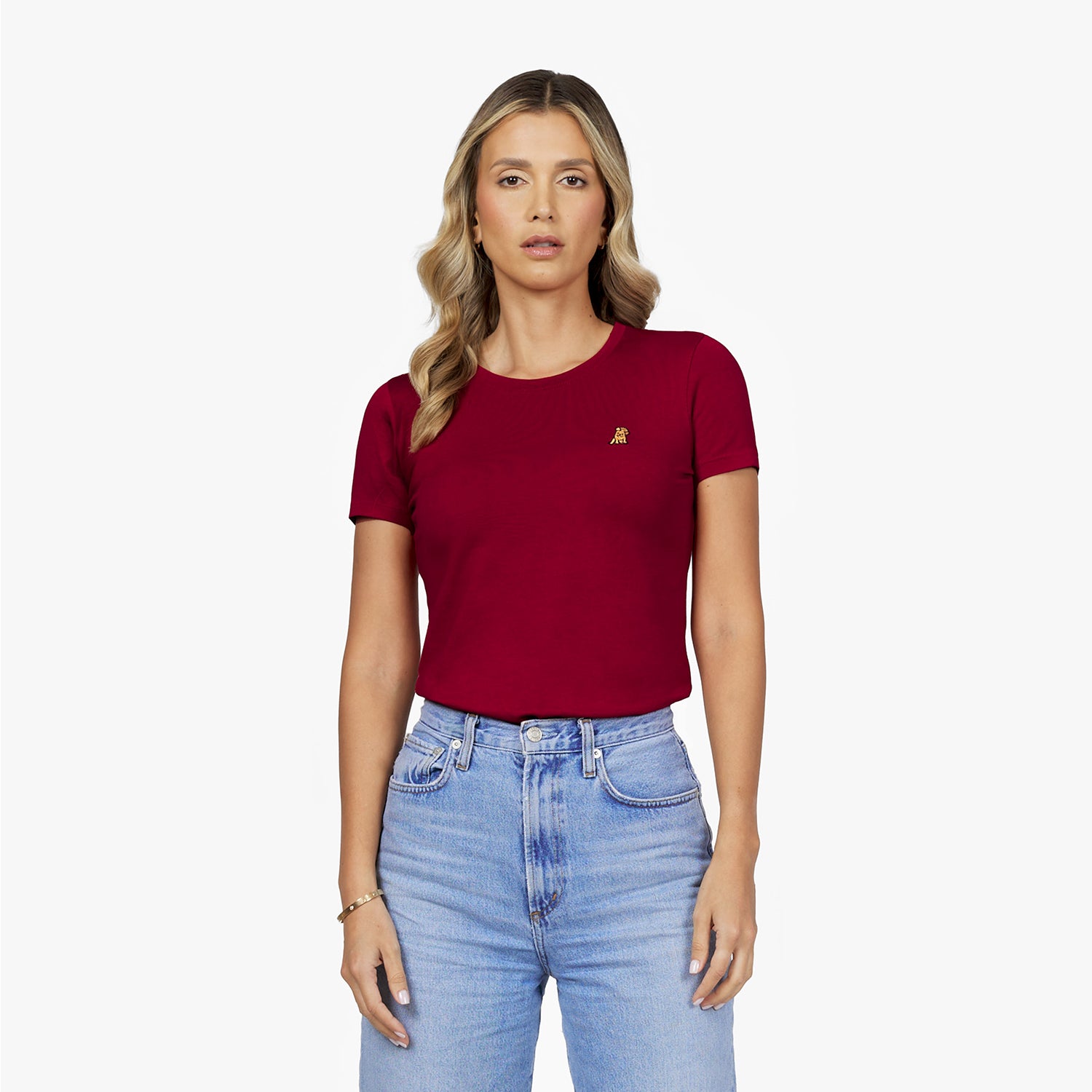 Front waist-up view of a female model wearing a burgundy crew neck t-shirt with a small gold embroidered logo on the left chest, paired with light wash jeans