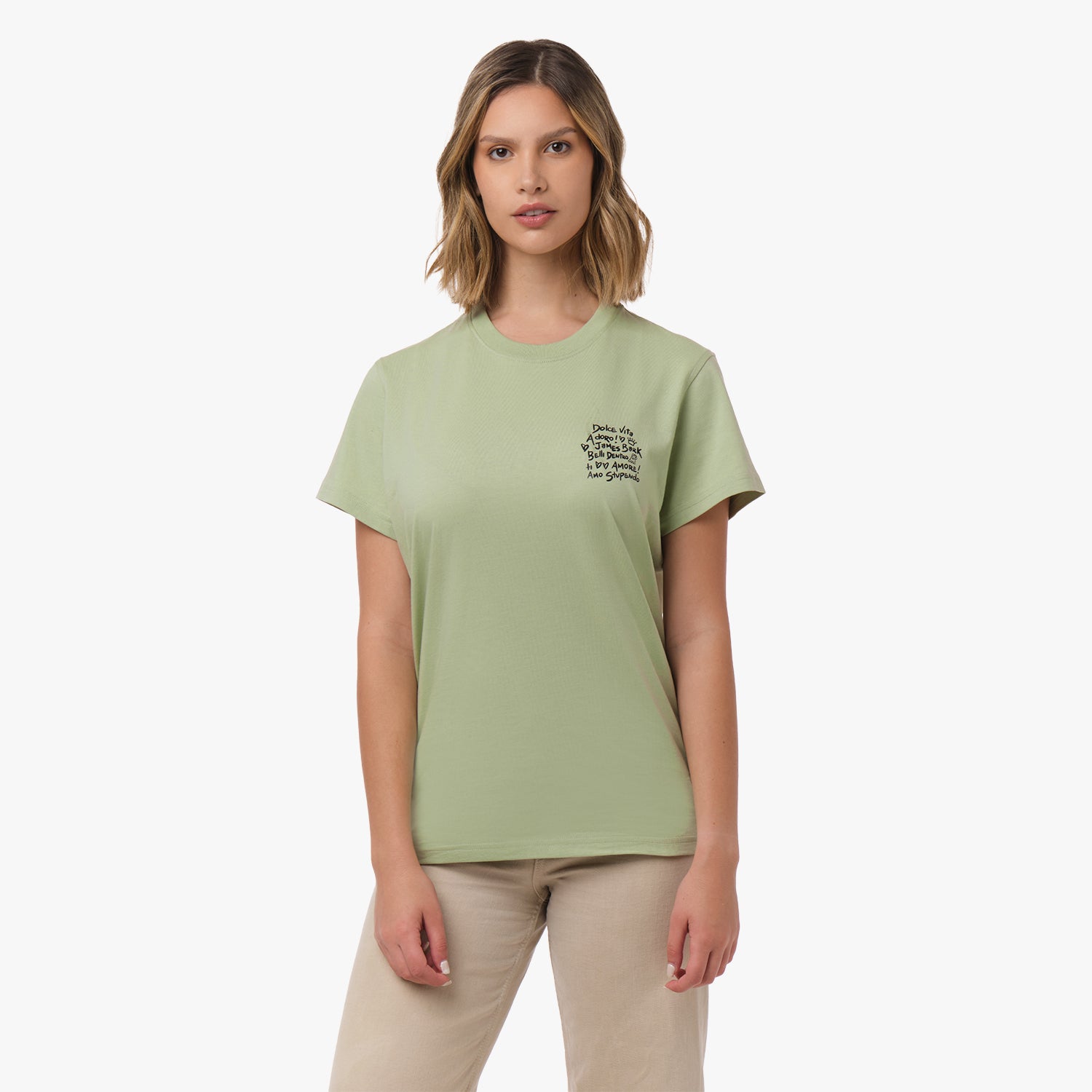 A woman stands facing forward, wearing a light green T-shirt with black text on the chest that includes phrases like "Dolce Vita," "Amore," and "Amo Stupendo." She pairs the shirt with beige pants and has a neutral expression.