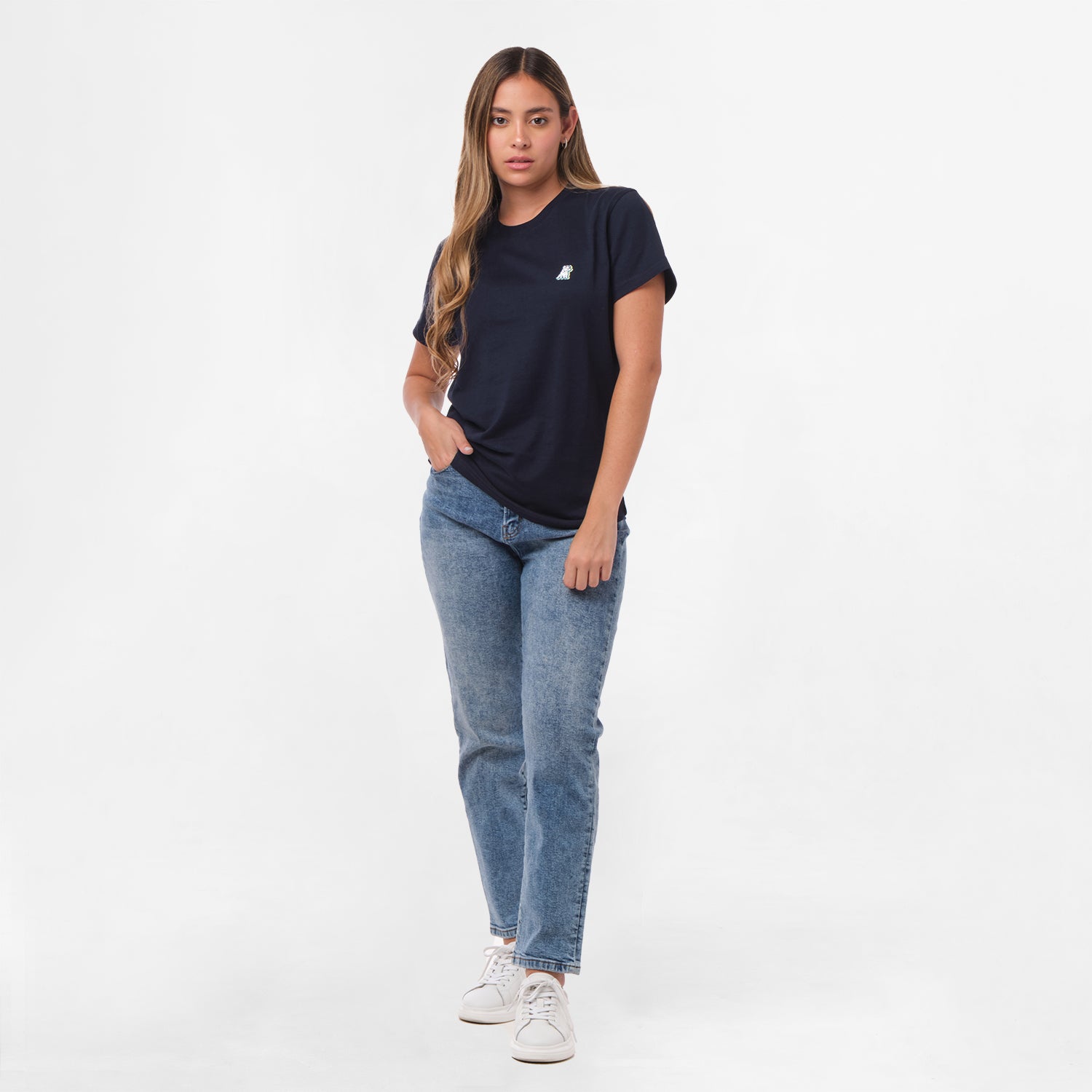 A full-body view of a woman wearing the navy blue T-shirt with the small embroidered dog logo on the chest, paired with blue jeans, standing with one hand in her pocket.