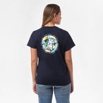 A woman is shown from behind, wearing a navy blue T-shirt with a large circular graphic on the back. The design features a dog sitting in front of a tropical background with the text "James Bark Privé Society" around the circle. She pairs the shirt with blue jeans.