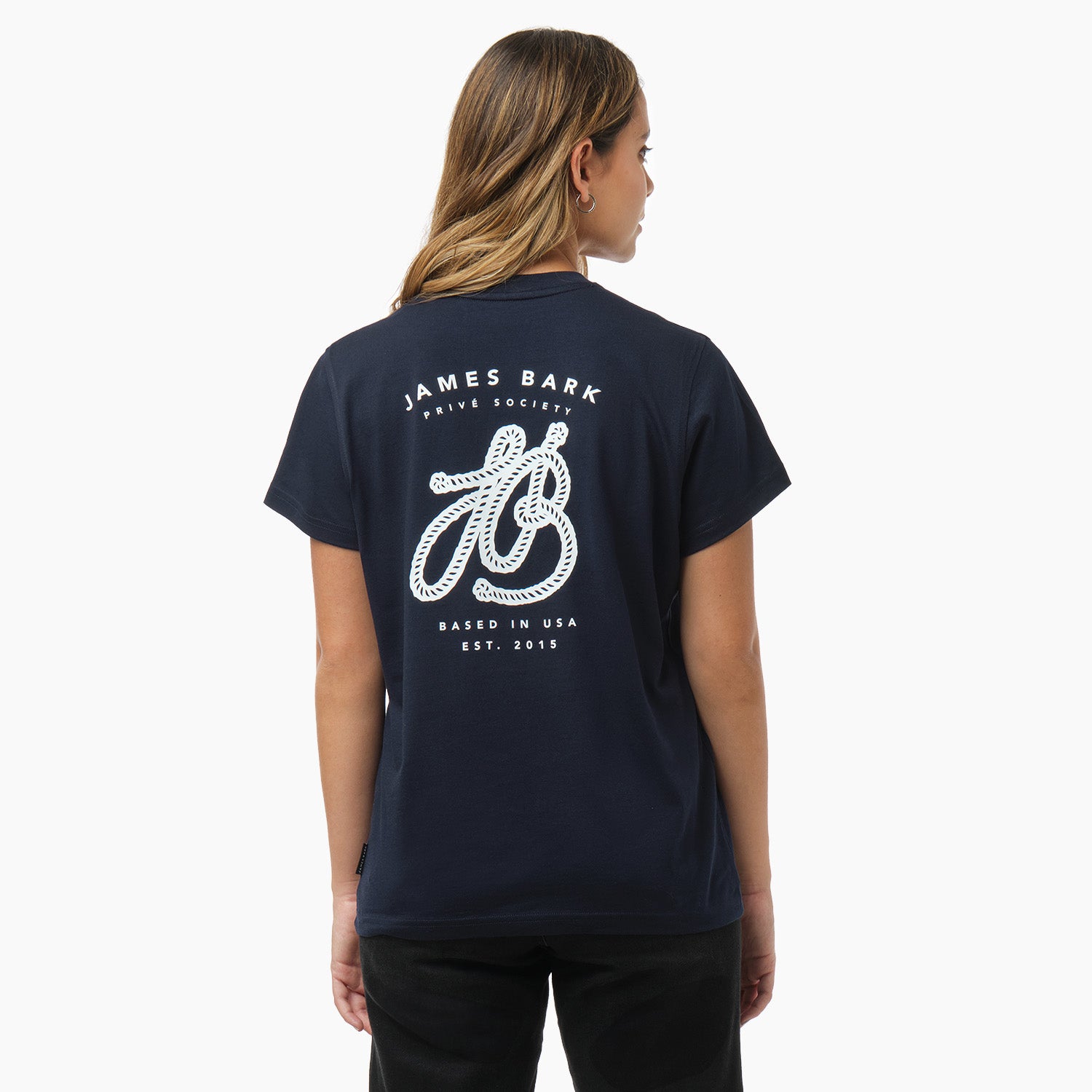 A woman is shown from behind, wearing a navy blue T-shirt with a large white graphic on the back. The design features the text "James Bark Privé Society," with a rope illustration forming the initials "JB" and the words "Based in USA, Est. 2015" below. She pairs the shirt with black pants.