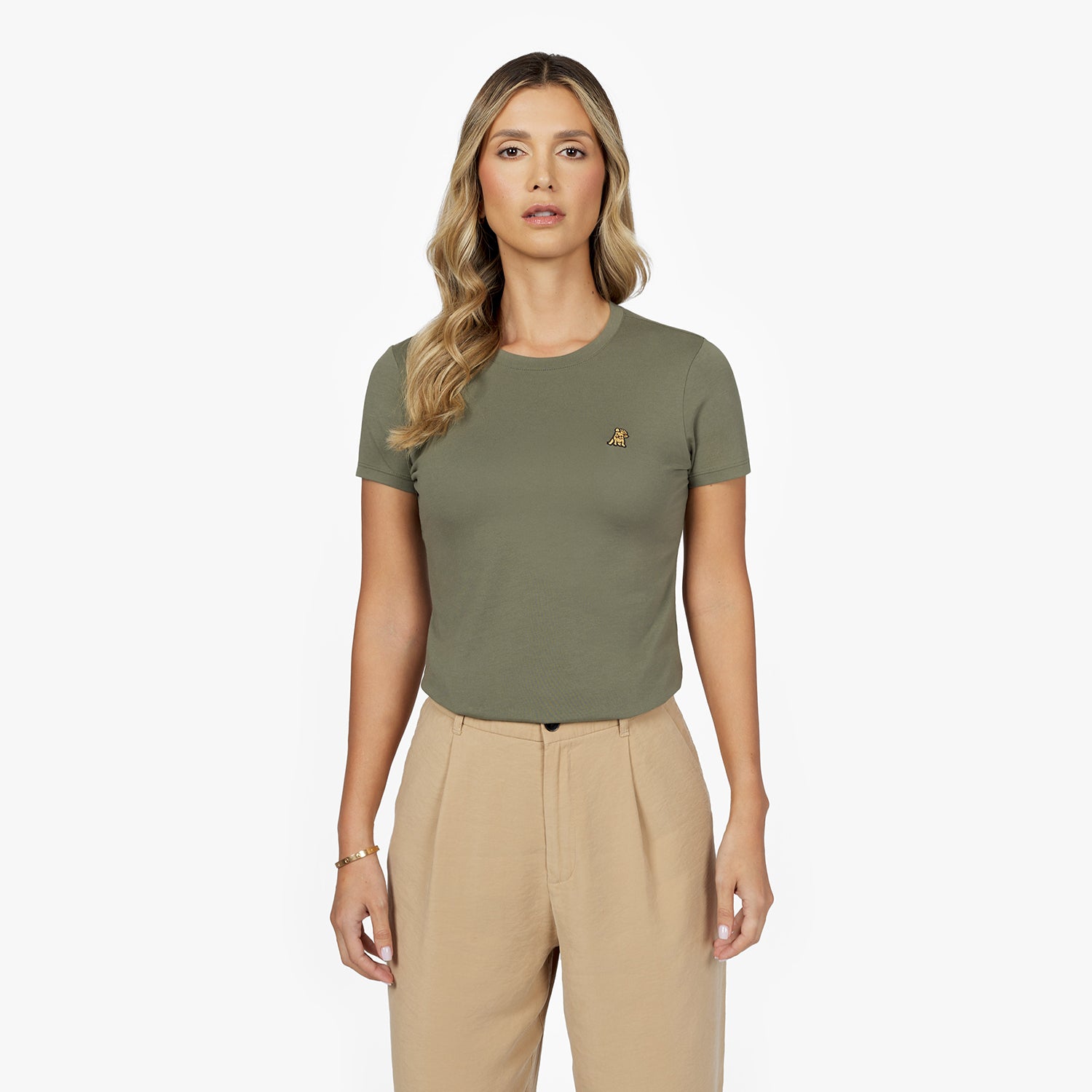 Front waist-up view of a female model wearing an olive green crew neck t-shirt with a small gold embroidered logo on the left chest, paired with beige trousers