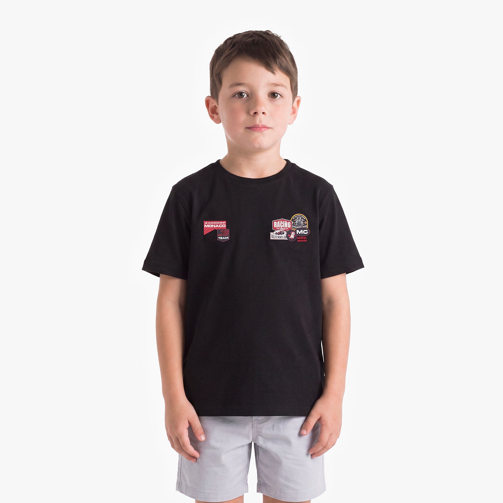 A young boy wearing a black T-shirt with two small racing-style chest patches and light gray shorts