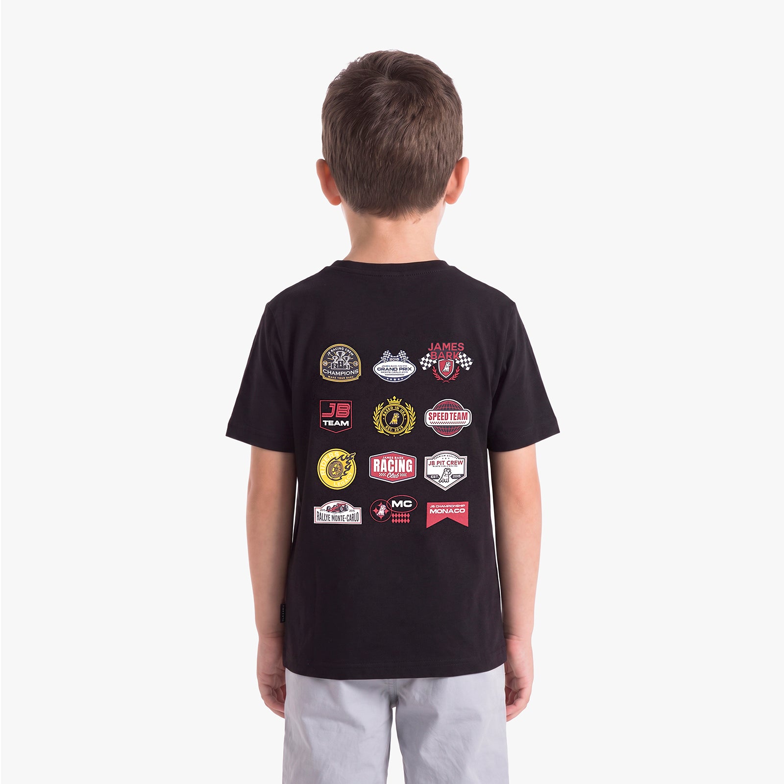 A child viewed from behind, wearing a black T-shirt with a collage of racing-themed patch graphics on the back