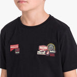 A close-up of a child wearing a black T-shirt with multiple racing-themed graphic patches on the chest, including Monaco, Racing, and Monte Carlo designs.