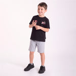 A young boy modeling a black T-shirt with a graphic patch on the left chest, paired with light gray shorts and black sneakers