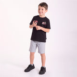 A young boy modeling a black T-shirt with a graphic patch on the left chest, paired with light gray shorts and black sneakers