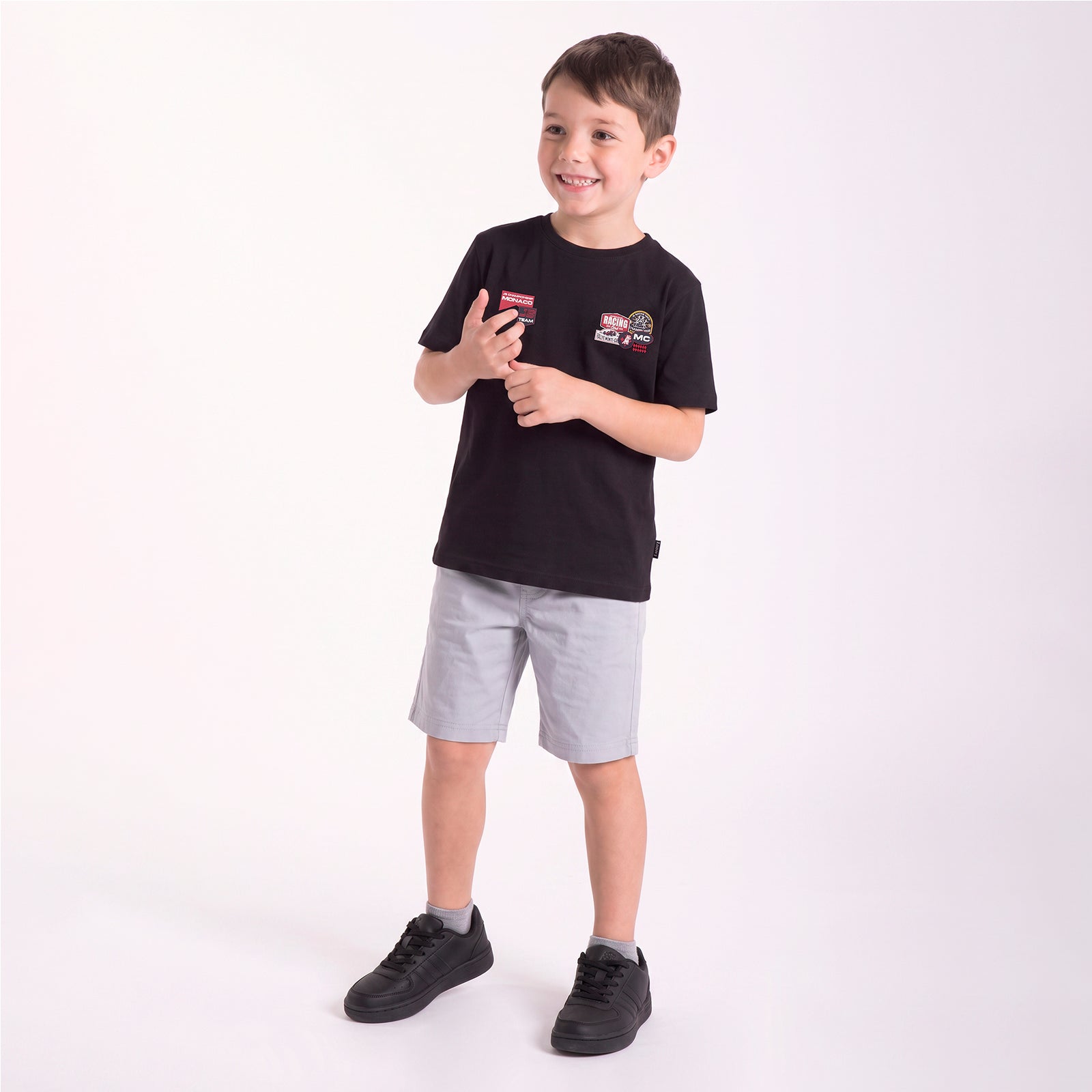 A young boy modeling a black T-shirt with a graphic patch on the left chest, paired with light gray shorts and black sneakers