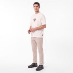A man modeling a light beige T-shirt with a small graphic on the left chest, paired with beige trousers and dark gray sneakers