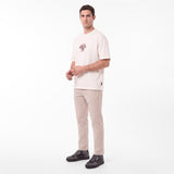A man modeling a light beige T-shirt with a small graphic on the left chest, paired with beige trousers and dark gray sneakers