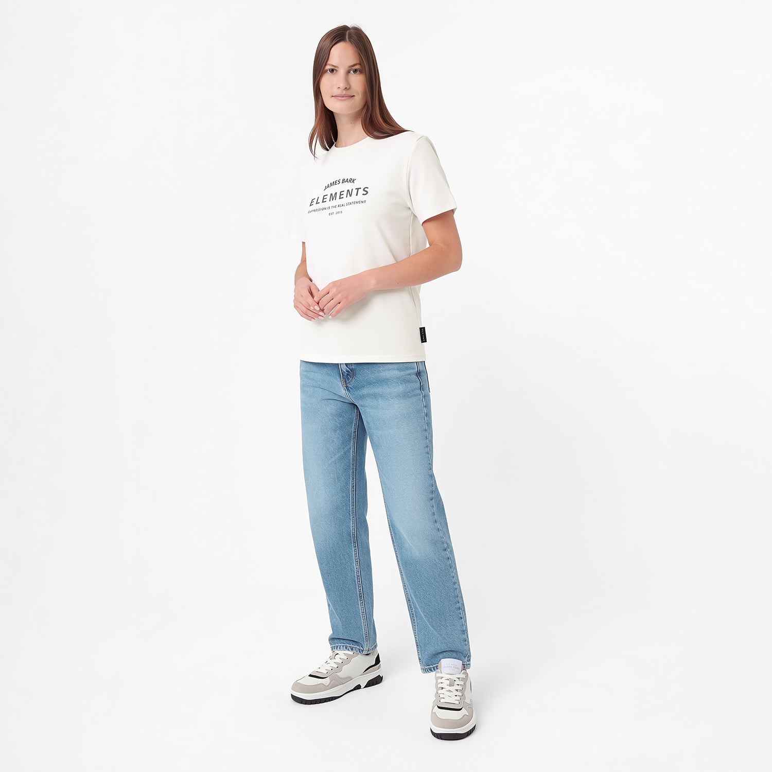 A woman modeling a white short-sleeve graphic T-shirt with 'ELEMENTS' printed on the chest, paired with light blue jeans and white sneakers