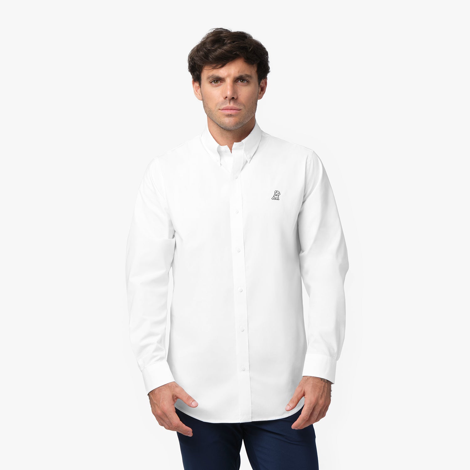 Full front view of a man wearing a white long-sleeved button-down shirt with a black and white logo and navy blue pants, standing against a white background