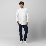 A man wearing a white long-sleeved button-down shirt and navy blue pants stands against a grey background with one hand in his pocket. He is also wearing white sneakers