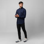 A man wearing a navy blue long-sleeved shirt, black pants, and sneakers stands against a grey background, looking left with his hands clasped