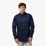 Full front view of a man wearing a navy blue long-sleeved button-down shirt with a white logo and light grey pants, standing against a white background