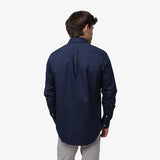 Back view of a man wearing a navy blue long-sleeved shirt and light grey pants against a white background, showing a box pleat on the back