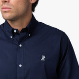 Close-up of a man wearing a navy blue long-sleeved button-down shirt, showing the collar, white buttons, and the embroidered white logo on the chest