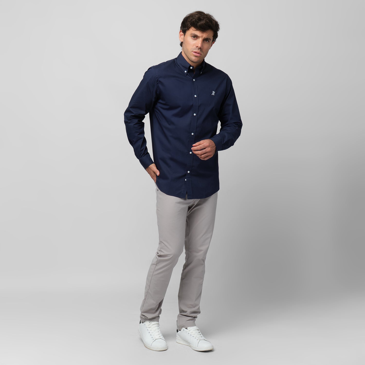 A man wearing a navy blue long-sleeved button-down shirt and light grey pants stands against a grey background, posing with one hand near his chest and the other in his pocket. He is also wearing white sneakers