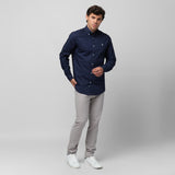 A man wearing a navy blue long-sleeved button-down shirt and light grey pants stands against a grey background, posing with one hand near his chest and the other in his pocket. He is also wearing white sneakers