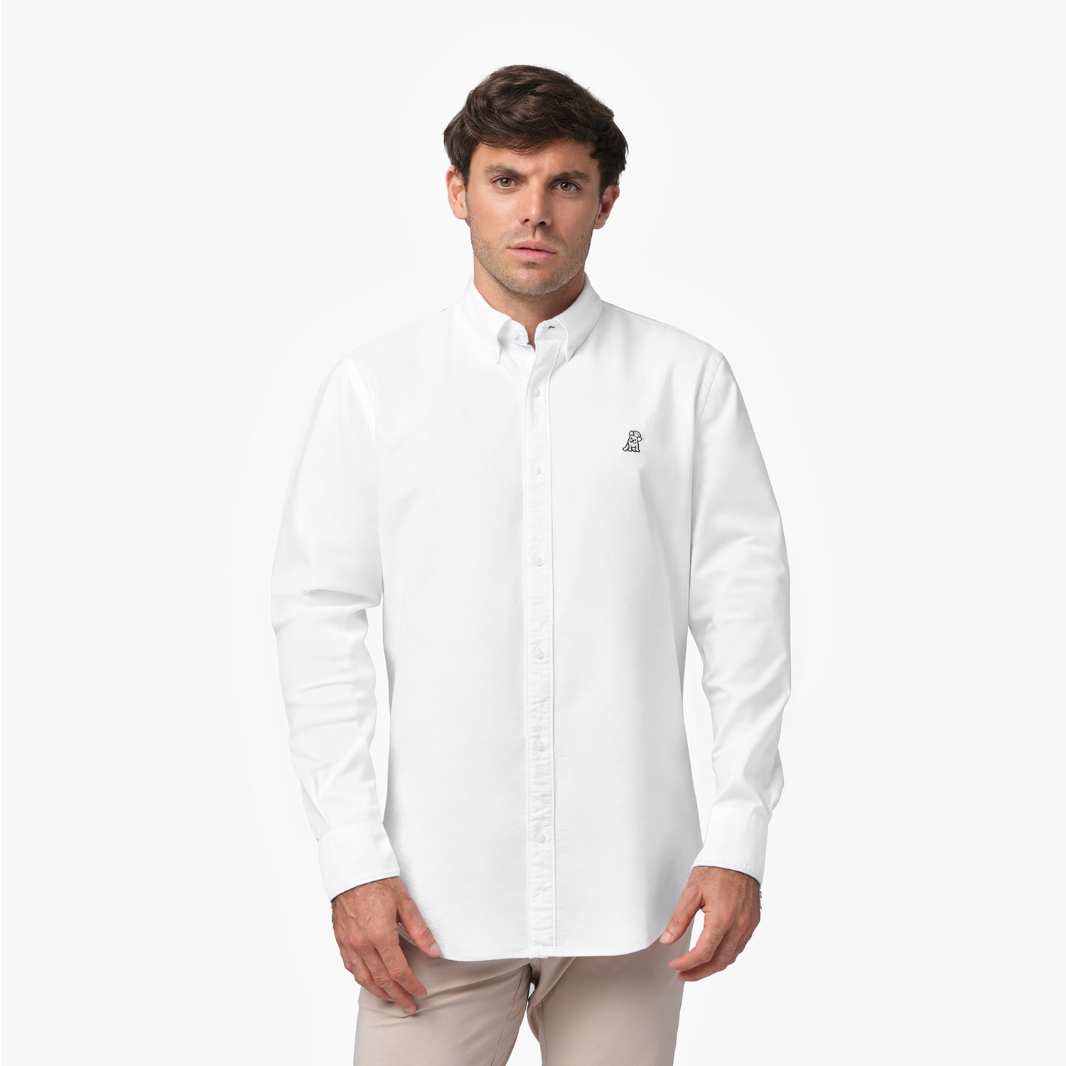Front waist-up view of a male model wearing a white long-sleeve button-down oxford shirt with a small black embroidered logo on the left chest, paired with beige trousers