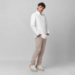 Full-length side/angled view of a male model wearing a white long-sleeve oxford shirt with a black logo, beige trousers, and white/beige sneakers, standing against a light grey background with hands in pockets