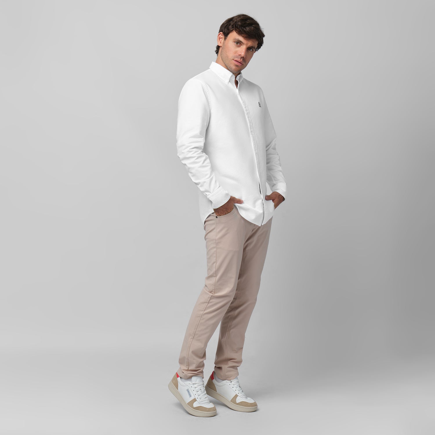 Full-length side/angled view of a male model wearing a white long-sleeve oxford shirt with a black logo, beige trousers, and white/beige sneakers, standing against a light grey background with hands in pockets