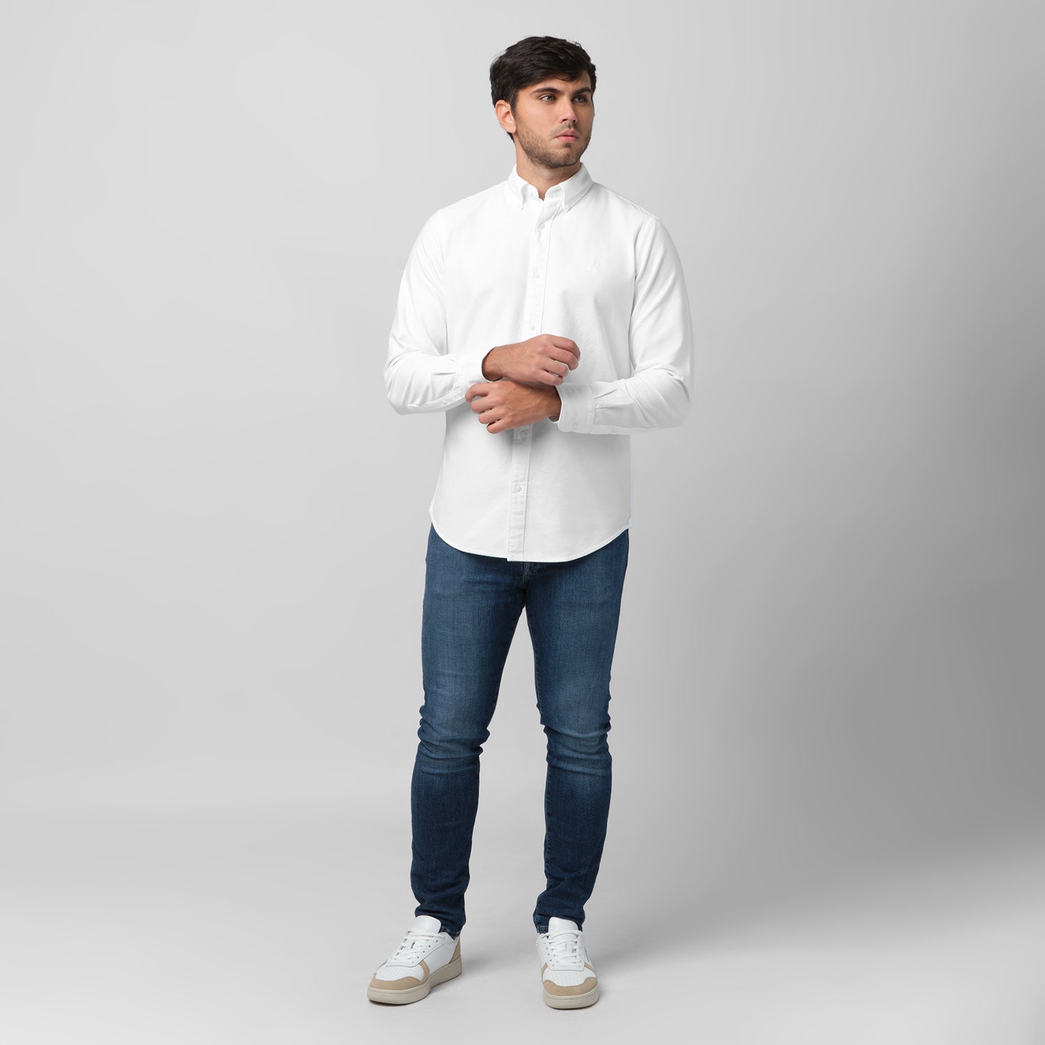 Full-length front view of a male model wearing a white long-sleeve oxford shirt with a white logo, dark wash jeans, and white/beige sneakers, standing against a light grey background while adjusting his cuff