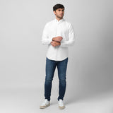 Full-length front view of a male model wearing a white long-sleeve oxford shirt with a white logo, dark wash jeans, and white/beige sneakers, standing against a light grey background while adjusting his cuff