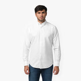 Front waist-up view of a male model wearing a white long-sleeve button-down oxford shirt with a small white embroidered logo on the left chest, paired with dark wash jeans