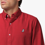 Close-up detail of the red oxford shirt collar and chest area on a male model, showing the button-down collar, buttons, fabric texture, and the small white embroidered dog logo on the left chest