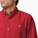 Close-up detail of the red oxford shirt collar and chest area on a male model, showing the button-down collar, buttons, fabric texture, and the small white embroidered dog logo on the left chest