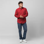 Full-length view of a male model wearing a red long-sleeve oxford shirt with a white logo, blue trousers, and white sneakers, standing against a light grey background with hands clasped