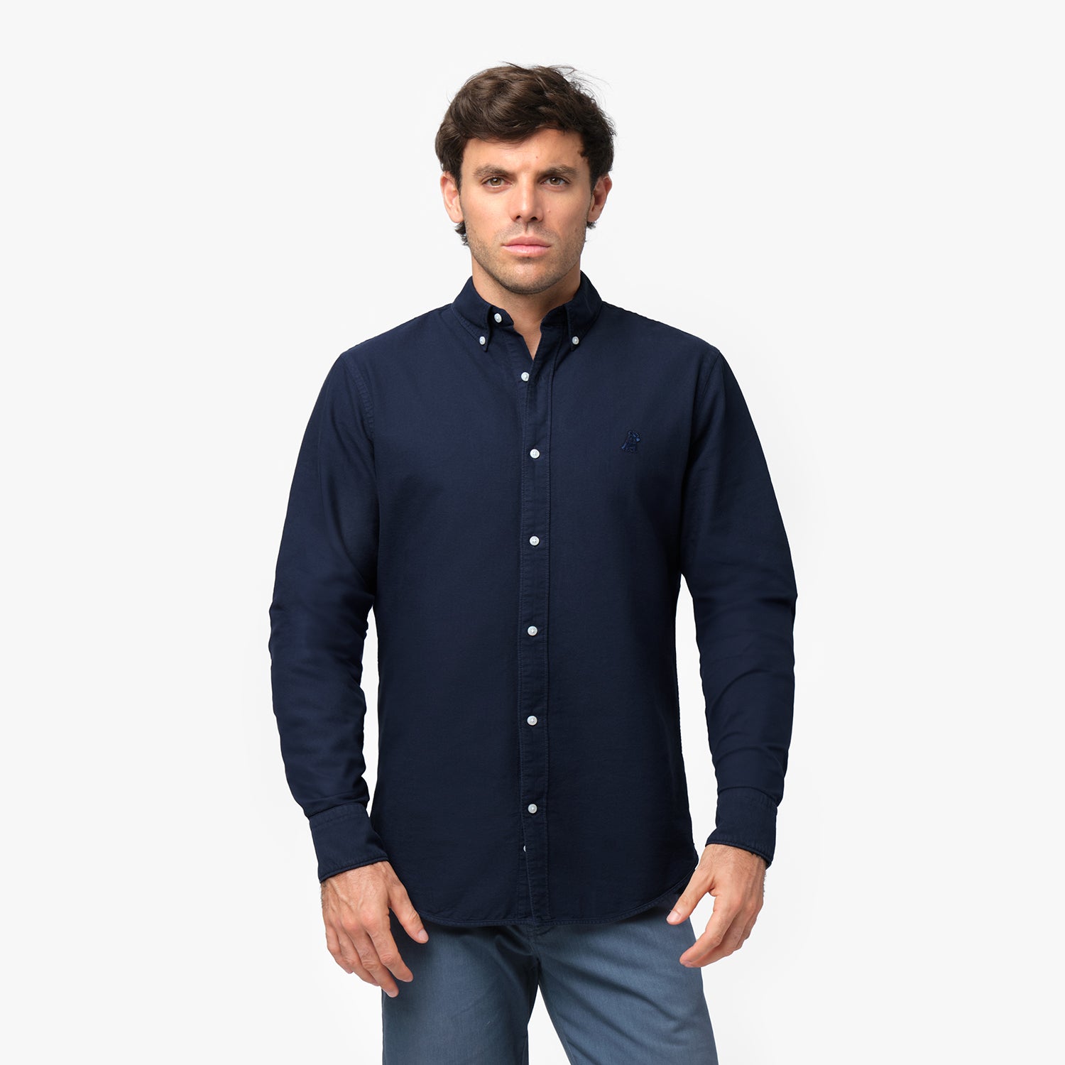 Front waist-up view of a male model wearing a navy blue long-sleeve button-down oxford shirt with a small blue embroidered logo on the left chest, paired with blue trousers