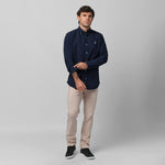 Full-length front view of a male model wearing a navy blue long-sleeve oxford shirt with a white logo, beige trousers, and black sneakers, standing against a light grey background while rolling up his sleeve