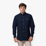 Front waist-up view of a male model wearing a navy blue long-sleeve button-down oxford shirt with a small white embroidered logo on the left chest, paired with beige trousers