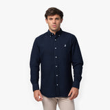 Front waist-up view of a male model wearing a navy blue long-sleeve button-down oxford shirt with a small white embroidered logo on the left chest, paired with beige trousers