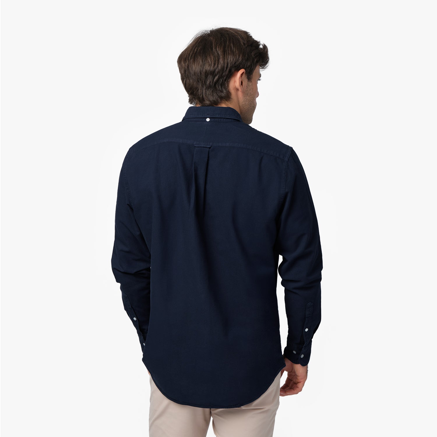 Back waist-up view of a male model wearing a navy blue long-sleeve oxford shirt with a back pleat, paired with beige trousers