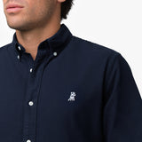 Close-up detail of the navy blue oxford shirt collar and chest area on a male model, showing the button-down collar, buttons, fabric texture, and the small white embroidered dog logo on the left chest