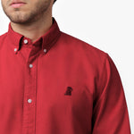 Close-up detail of the red oxford shirt collar and chest area on a male model, showing the button-down collar, buttons, fabric texture, and the small black embroidered dog logo on the left chest