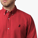 Close-up detail of the red oxford shirt collar and chest area on a male model, showing the button-down collar, buttons, fabric texture, and the small black embroidered dog logo on the left chest