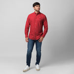 Full-length angled view of a male model wearing a red long-sleeve oxford shirt with a black logo, dark wash jeans, and white/beige sneakers, standing against a light grey background with one hand in his pocket