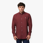 Front waist-up view of a male model wearing a burgundy long-sleeve button-down oxford shirt with a small white embroidered logo on the left chest, paired with navy trousers