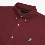 Close-up of the collar and upper chest area of a maroon button-down shirt. Features include a button-down collar, white buttons, a black "JAMES BARK" brand tag, and a black-and-white embroidered dog logo on the left chest