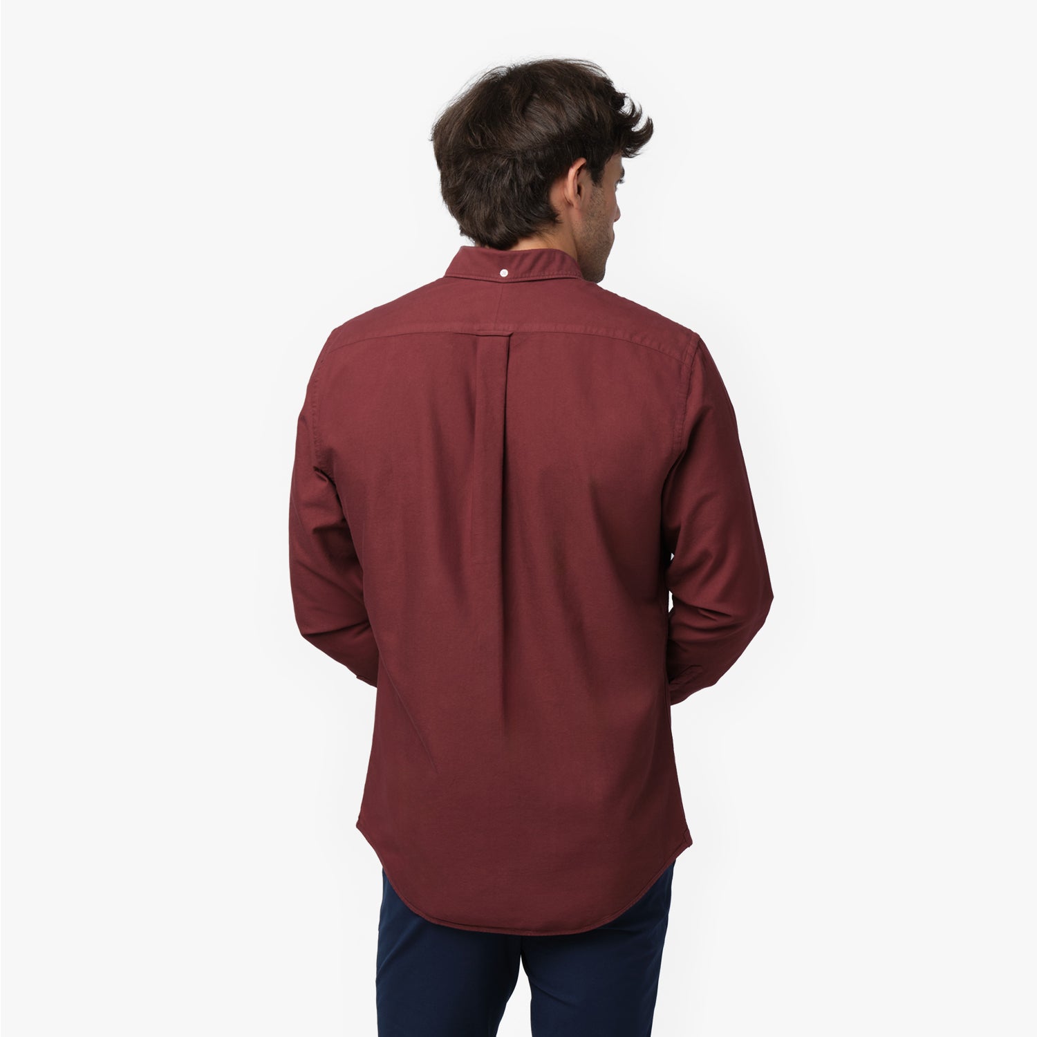 Back waist-up view of a male model wearing a burgundy long-sleeve oxford shirt with a back pleat, paired with navy trousers