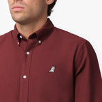Close-up detail of the burgundy oxford shirt collar and chest area on a male model, showing the button-down collar, buttons, fabric texture, and the small white embroidered dog logo on the left chest